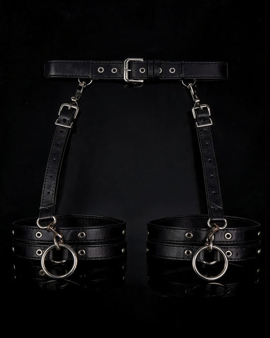Dominatrix black faux-leather harness belt with thigh cuffs and silver O-rings, black background